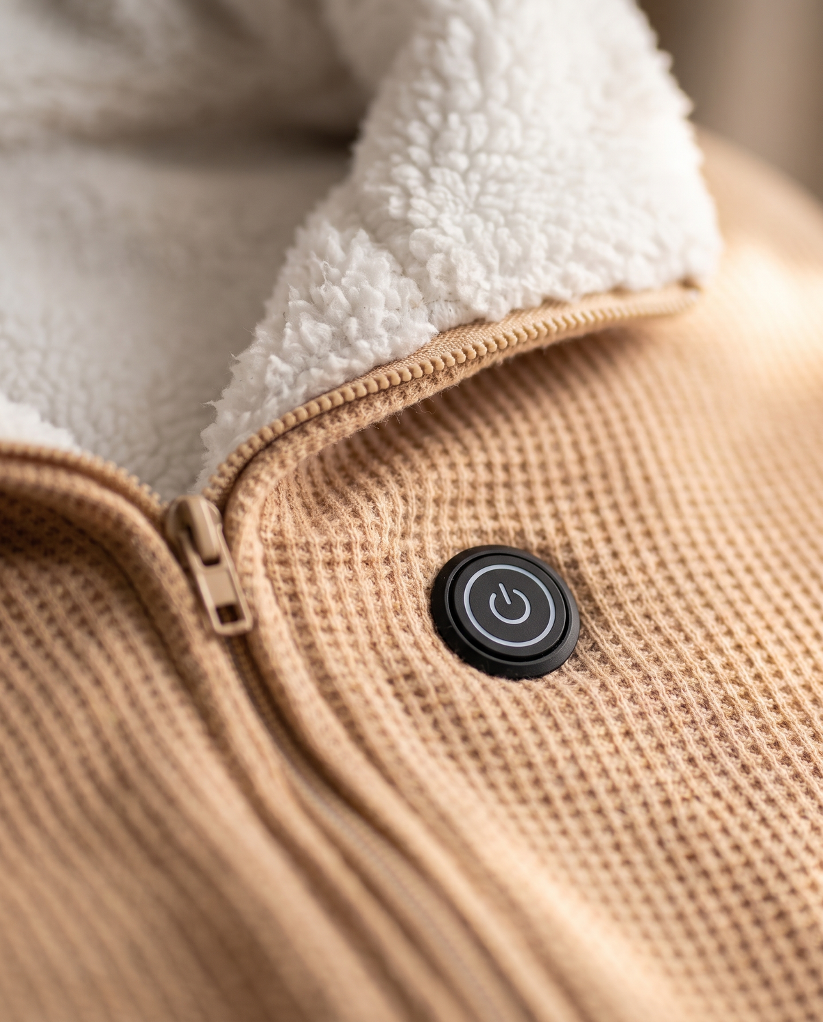 Close-up of the heated shawl's one-touch power button on waffle-knit fabric beside the zip