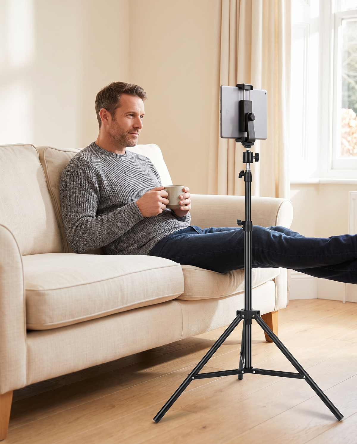 Man relaxing on a sofa with a mug, watching a tablet held at eye level by the tripod floor stand